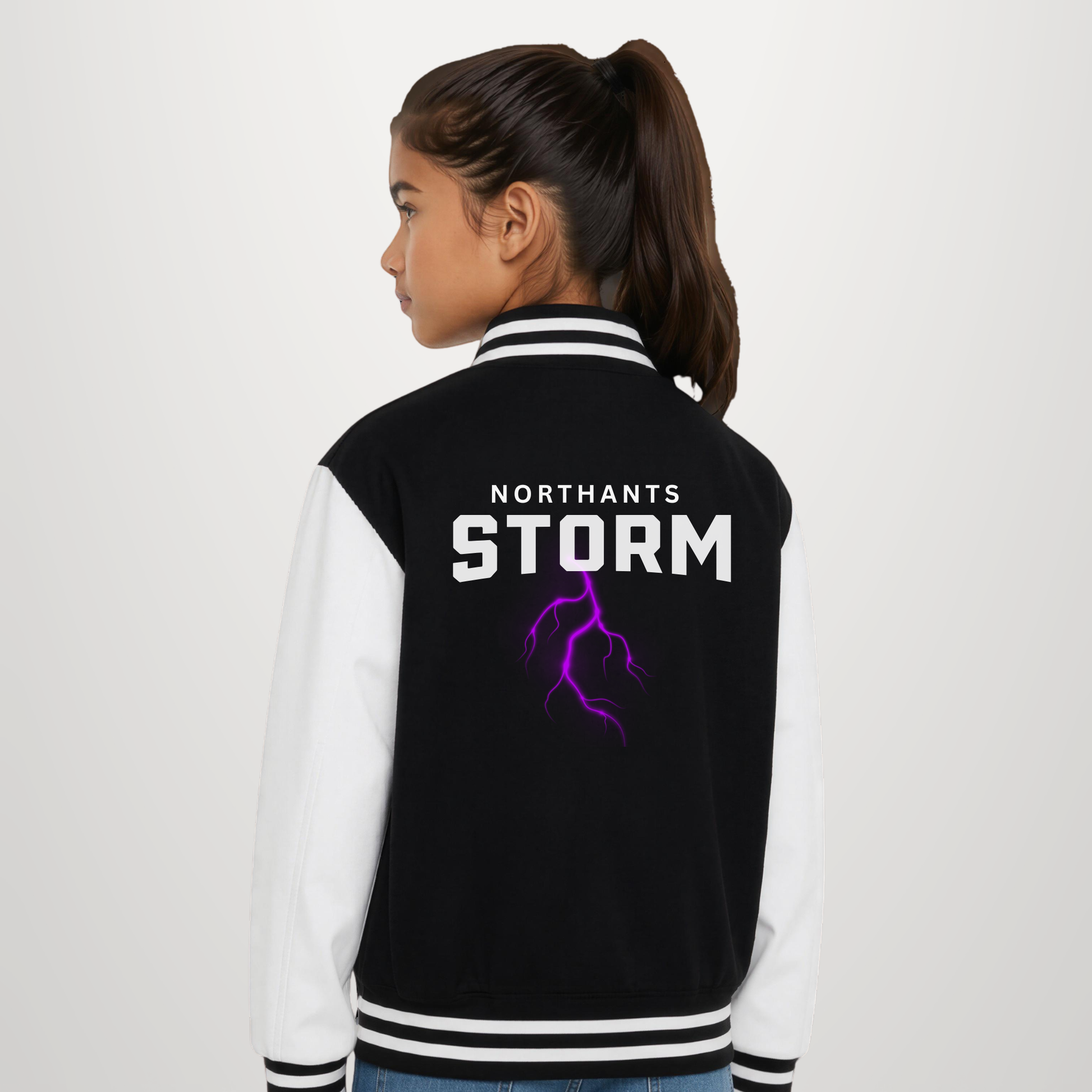 A girl shown from the back wearing a varsity-style jacket with black body panels, white sleeves, and a purple lightning graphic with the text 'NORTHANTS STORM' on the back.