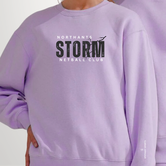 A person wearing a lavender crewneck sweatshirt with a black and white graphic on the chest that reads 'NORTHANTS STORM NETBALL CLUB'.