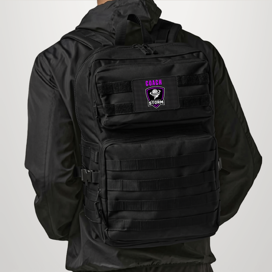 A black tactical backpack worn by a model, viewed from the back. The backpack features a front zippered pocket with a black velcro patch displaying the text 'COACH STORM' in purple and white.