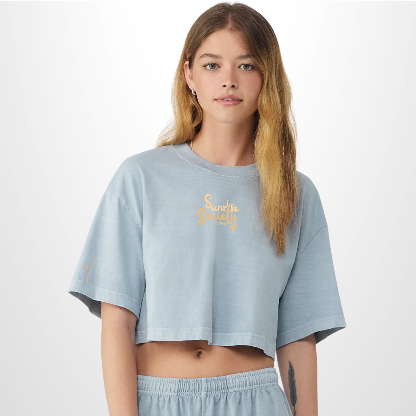 A woman modeling a light blue cropped t-shirt with a yellow 'Sunrise Society' logo on the chest, paired with matching light blue sweatpants, against a plain white studio background.